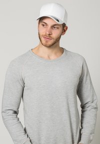 Grijze sweatshirt met raglanmouwen, lichte stikselaccenten en een ronde halslijn. Gecombineerd met een effen witte pet.