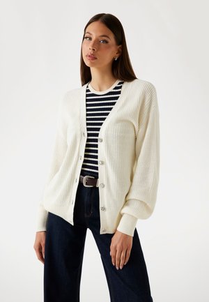 Cardigan lavorato a maglia di colore crema con una vestibilità ampia, dotato di grandi bottoni e polsini a coste, indossato sopra una camicia a righe blu navy e bianca e pantaloni scuri.