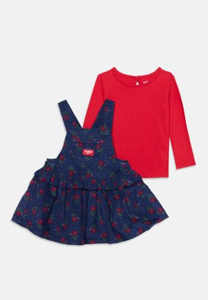 Tenue pour enfant composée d’un haut rouge à manches longues et d’une robe chasuble bleu marine à fleurs, avec des bretelles et une jupe à volants.