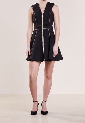 Kobieta ubrana w czarną mini sukienkę bez rękawów z złotym zamkiem błyskawicznym i obszyciami, do tego czarne wysokie obcasy na kostce na neutralnym tle.