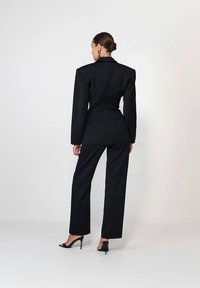 Femeie cu părul prins în coc, îmbrăcată într-un sacou negru cu umeri evidențiați, pantaloni asortați și sandale cu toc înalt negre, stând pe un podea albă.