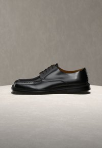 Μαύρο γυαλιστερό δερμάτινο παπούτσι με κορδόνια και λεπτομερείς ραφές, σε ανοιχτή επιφάνεια με ουδέτερο φόντο.