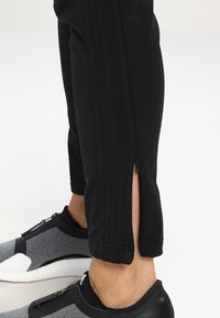 Dolní část nohy v černých kalhotách s otevřeným bočním zipem, spárovaná s černými a šedými slip-on sportovními botami na bílém pozadí.