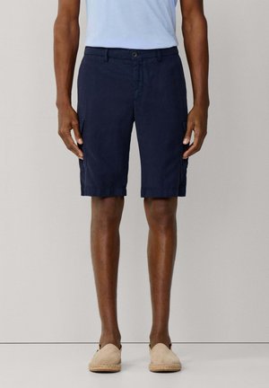 Hombre que lleva pantalones cortos cargo azul marino hasta la rodilla, camiseta azul claro y zapatos beige sin cordones, de pie contra un fondo neutro.