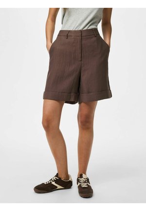 Vrouw draagt bruine omgeslagen korte broek, grijs T-shirt en bruine sneakers met witte veters, staat met handen in de zakken tegen een witte achtergrond.