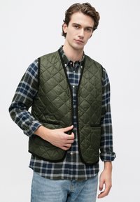 Νεαρός άνδρας φορεί ένα πράσινο καπιτονέ γιλέκο πάνω από ένα καρό πουκάμισο σε μπλε και γκρι αποχρώσεις με ανοιχτό μπλε τζιν, προσαρμόζοντας το γιλέκο με το ένα χέρι.