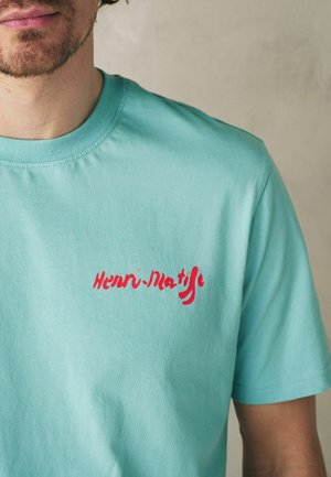 Man wearing light turquoise T-shirt with red "Henri-Matisse" text printed on the left chest against a plain beige background.