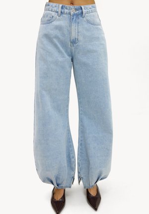 Jean large taille haute bleu clair avec ourlets froncés et fentes aux chevilles, porté avec des chaussures pointues marron foncé.