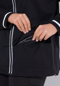 Czarna kurtka z białymi akcentami, z kieszenią na zamek i elastycznymi mankietami. Materiał wydaje się gładki o matowym wykończeniu.