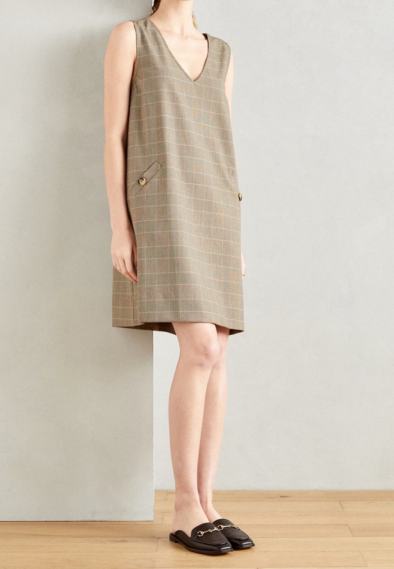 Femme portant une robe beige à carreaux sans manches avec deux boutons sur le devant et des mocassins marron foncé sans lacets, debout sur un sol en bois.