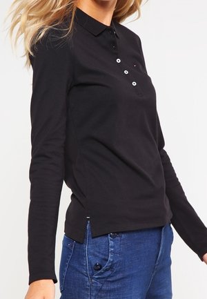 Femme portant un polo noir à manches longues et un jean bleu, posant avec les cheveux flottant sur le côté devant un fond uni.