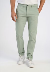 Chinos vert clair en tissu coton mélangé, avec une coupe droite, des poches frontales et une fermeture à un seul bouton. Assortis à des baskets blanches.