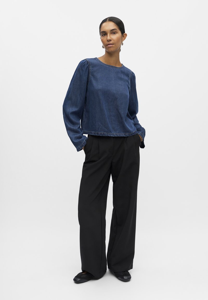 Tamni traper krojeni top s dugim rukavima, okruglim izrezom i strganim rubom. Uparen s labavim crnim hlačama i crnim ravnim cipelama.