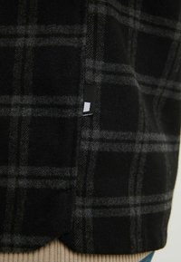 Gros plan sur un tissu à carreaux noir et gris avec une petite étiquette en tissu noire et blanche sur la couture latérale.