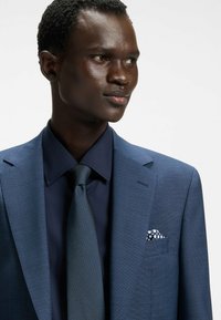 Blå texturerad kavaj med notch-öppningar, i kombination med en mörkblå skjorta och slips. Fickfyrkant har ett vitt prickmönster.