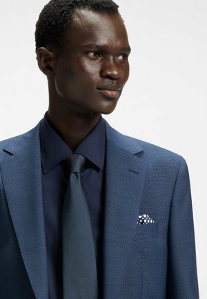 Veste de costume texturée bleue avec revers à cran, associée à une chemise et une cravate bleu foncé. Le pochettte a un motif à pois blancs.