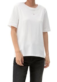 Biała bawełniana koszulka z krótkim rękawem, okrągłym dekoltem i małym haftowanym okręgiem na klatce piersiowej. Połączona z czarnymi dżinsami.