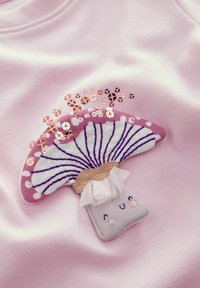 Un tissu rose clair présente un graphique de champignon avec un chapeau texturé, des détails brodés et des paillettes, accentué par un élément en vagues blanc.