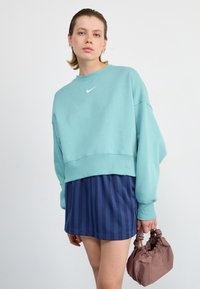 Türkiz Nike pulóver, rövidített fazonban, tengerészkék csíkos szoknyával és barna textúrájú kézitáskával párosítva. Sima anyag, laza stílus.