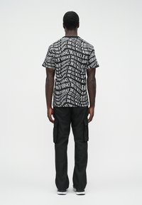 Muž otočený chrbtom, oblečený v čierno-bielej grafickej tričke s textom "Versace Jeans Couture", čiernych cargo nohaviciach s vreckami a bielych teniskách.