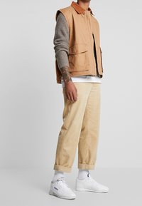 Tan bomull utanärmad jacka med brun kordflorskrage över en grå långärmad tröja, tillsammans med beige kordflorsbyxor och vita sneakers.
