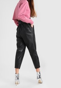 Mujer con pantalones de cuero negros de cintura alta, suéter corto rosa y botas de tacón alto metálicas plateadas, de pie contra un fondo blanco.