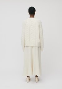 Un suéter de punto trenzado color crema con mangas largas, combinado con una falda larga a juego. El conjunto presenta un corte holgado y una textura suave, complementado por unos tacones beige.