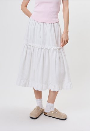 Femme portant une jupe midi blanche à volants, un haut rose sans manches, des chaussettes blanches et des chaussures beiges sans lacets.