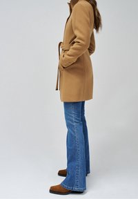 Cappotto di lana color cammello con cintura annodata, caratterizzato da un collo alto e polsini con bottoni, abbinato a jeans svasati blu e stivaletti marroni.