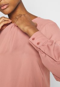 Maglietta rosa a maniche lunghe con scollatura arricciata e dettagli con polsini bottonati, realizzata in tessuto leggero e con una texture liscia.