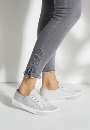 Baskets en toile grises à enfiler avec des semelles en caoutchouc blanches, dotées d'œillets en métal. Portées avec un jean skinny gris, montrant un détail de laçage au niveau de la cheville.