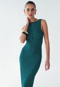 Žalsvai mėlyna, tekstūruota suknelė, aptempianti figūrą ir be rankovių. Papildyta apskritu iškirpimu ir surinktų raukinių juosmens detalėmis.