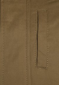 Materiał khaki o gładkiej fakturze, z dwoma pionowymi szwami i małą kieszonką naszytą po lewej stronie. Delikatne złote przeszycia.