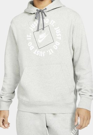 Osoba nosząca jasnoszarą bluzę z kapturem Nike i pasujące spodnie dresowe z napisem "Just Do It" i logo Nike na piersi.