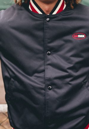 Osoba ubrana w czarną zapinaną na guziki kurtkę z czerwono-białymi paskami na kołnierzu oraz małym czerwonym logo "air max" na piersi.