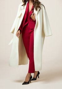 Manteau en laine blanche, costume rouge sur mesure avec blouse et pantalon, chaussures noires à talons hauts. Texture lisse et design structuré.