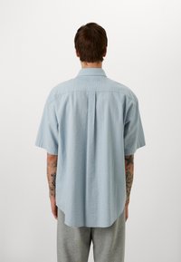 Chemise bleu clair à manches courtes avec un motif texturé et un col à boutons, présentant un pli au dos et un ourlet légèrement arrondi.
