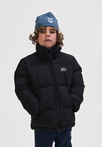 Melna pūkaina jaka ar augstu apkakli, priekšpusē rāvējslēdzēja aizdare un neliels logotipa plāksteris. Valkātājam ir cirtaini mati un zilā cepure.