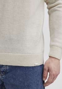 Jersey de punto en crema claro con puños y dobladillo acanalados. Etiqueta en el dobladillo inferior. Combinado con jeans de mezclilla azules. Textura suave, diseño minimalista.