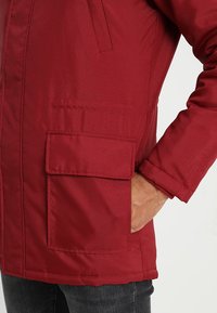 Chaqueta aislante roja con cremallera frontal, bolsillo lateral y tela texturizada. Cuenta con cuello alto y mangas ligeramente entalladas.