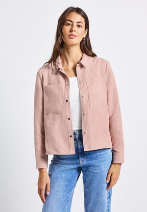 Femme portant une veste en velours côtelé rose clair à boutons sur un top blanc et un jean bleu, se tenant devant un fond blanc.