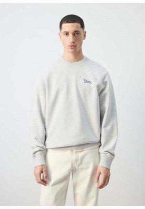 Młody mężczyzna z krótkimi ciemnymi włosami, ubrany w jasnoszarą bluzę z napisem „Yes.” wyszytym na piersi niebieską nicią oraz kremowe spodnie.