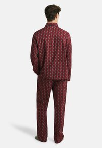 Man standing facing away wearing matching red paisley patterned pajama shirt and pants with gray slippers on white background.