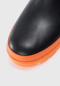 Botte en caoutchouc noir avec une semelle orange texturée. L'embout présente un design lisse et arrondi, et la semelle est dotée d'un motif pour une meilleure adhérence.