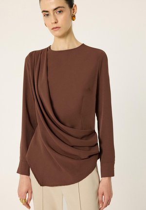 Femme portant un haut marron à manches longues avec un tissu drapé sur un côté et un pantalon beige, accessoirisée avec des boucles d’oreilles en ambre et une bague en or.