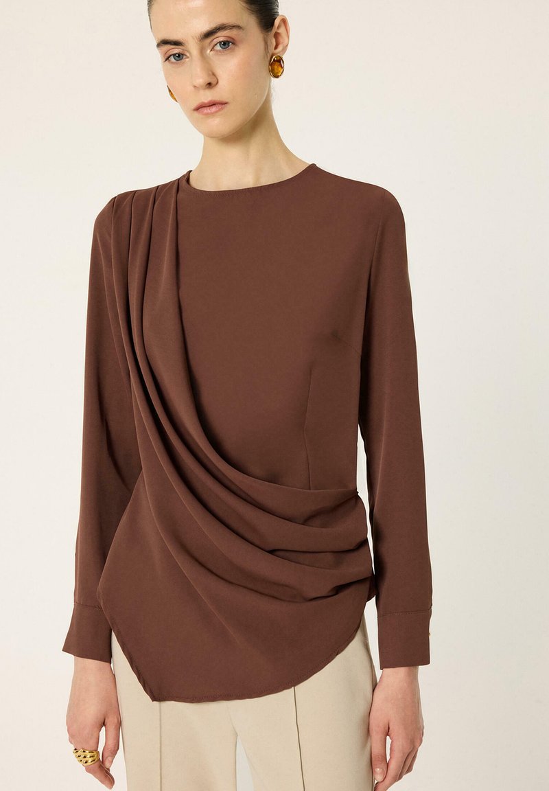 Femme portant un haut marron à manches longues avec un tissu drapé sur un côté et un pantalon beige, accessoirisée avec des boucles d’oreilles en ambre et une bague en or.