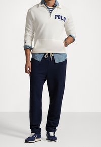 Man med vit Polo Ralph Lauren-tröja över en blå chambrayskjorta, marinblå joggingbyxor och blå sneakers med vita snören och ränder.