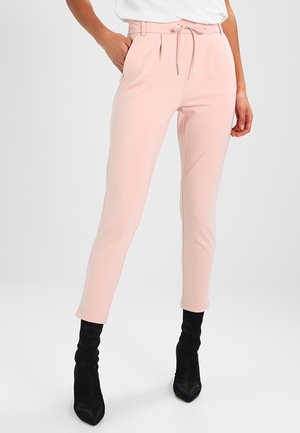 Vrouw met een lichtroze, hoogwater, taps toelopende broek met trekkoord in de taille en zwarte pumps met hoge hak en spits toe, staand met één hand in de zak.