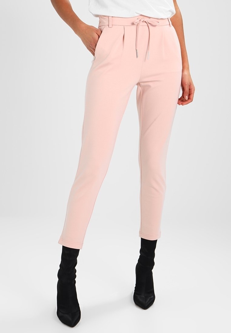 Femme portant un pantalon taille haute fuselé rose clair avec une taille à cordon et des bottines noires à talons pointues, debout avec une main dans la poche.
