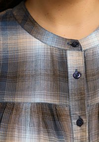 Ruudullinen nappipaita sinisessä, ruskeassa ja kermassa. Ominaisuuksiin kuuluu pyöreä kaulus, kaksi nappia ja teksturoitu kangas pehmeällä pinnalla.